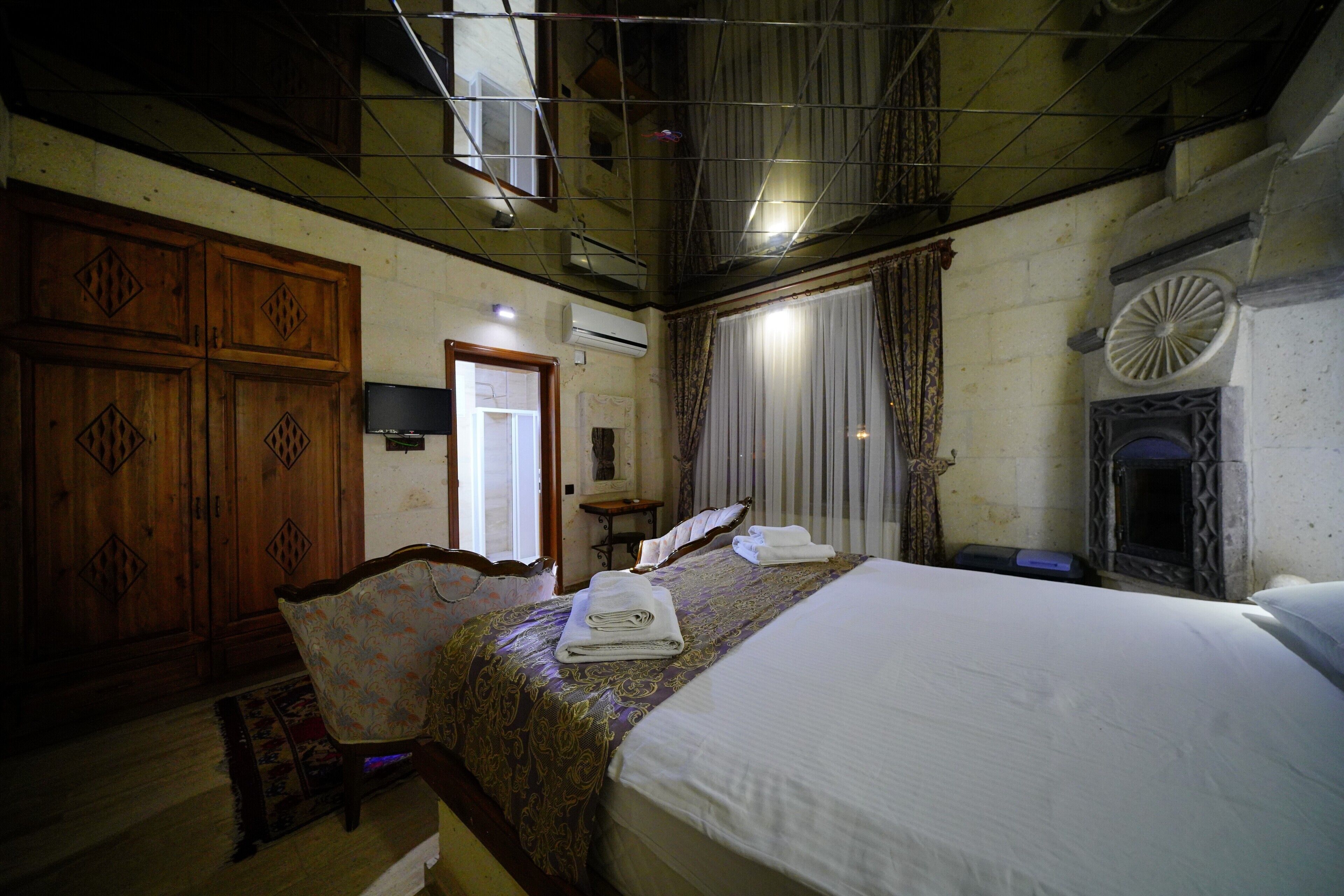 A hotel room with a large bed, a wooden wardrobe, a small table, and a fireplace.