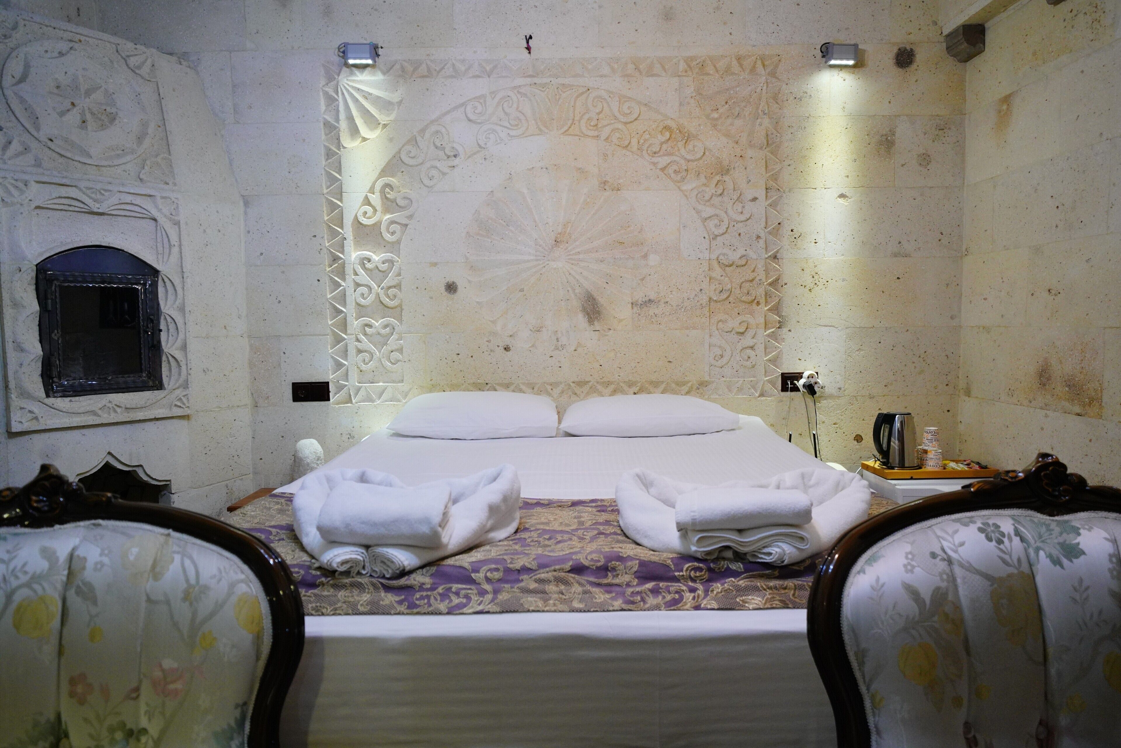 A hotel room with a large bed, two armchairs, a decorative wall, and a small table with a kettle.