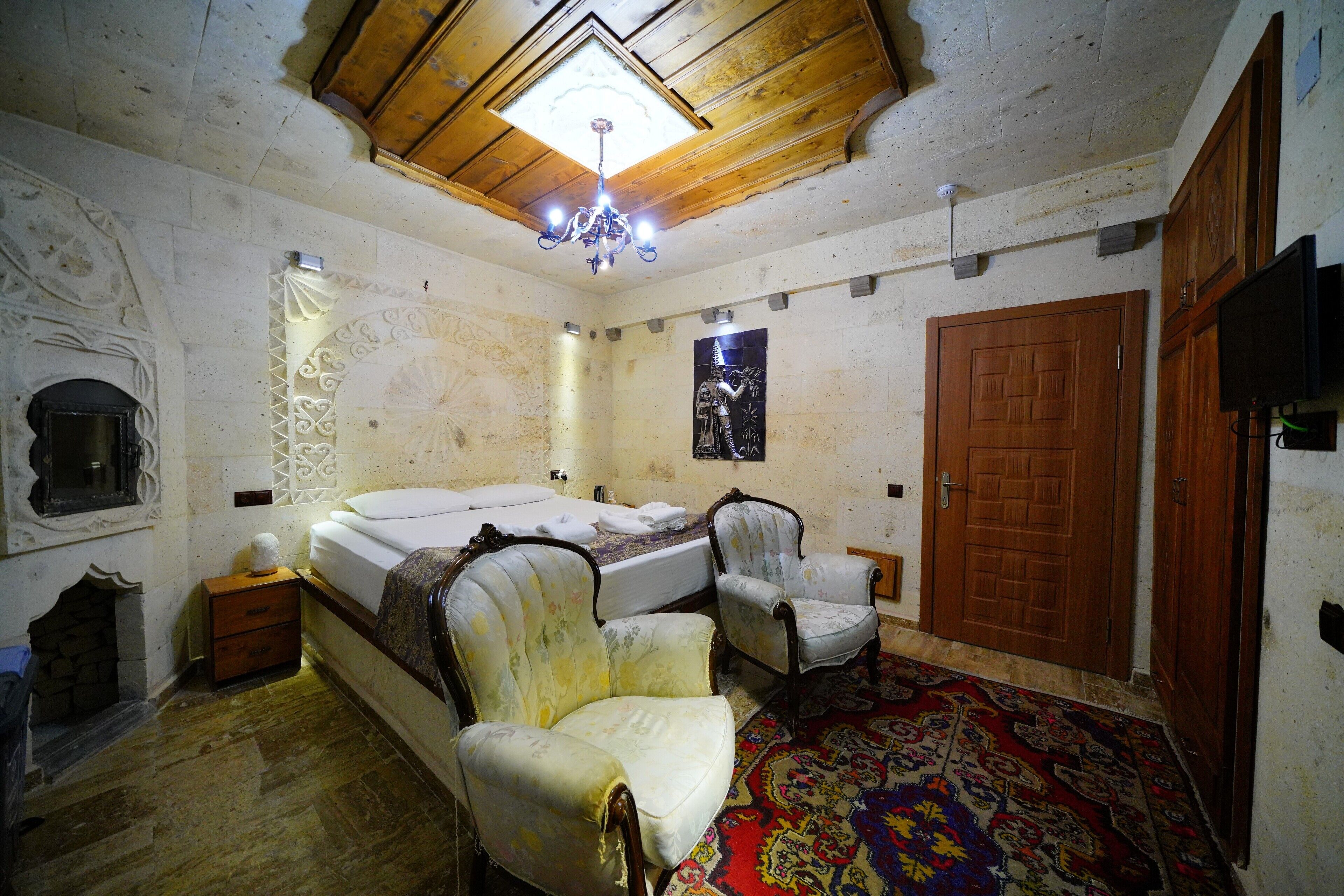 Un dormitorio con una cama, dos sillas, techo de madera, una araña, un tapete estampado y una puerta de madera.