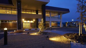 Un edificio moderno con una entrada bien iluminada, ventanas amplias y un área ajardinada con un árbol pequeño y defensas antivallas.