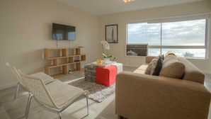 A modern living room with a large window, a flat-screen TV, a wooden bookshelf, and a red ottoman. - Vue Devant (Cape Town)