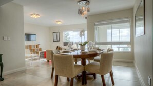 A dining area with a round table, chairs, and a chandelier. There is a TV mounted on the wall and a window providing natural light. - Vue Devant (Cape Town)