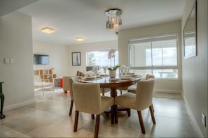A dining area with a round table, chairs, and a chandelier. There is a TV mounted on the wall and a window providing natural light. - Vue Devant (Cape Town)