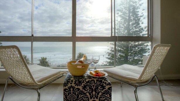 A room with two wicker chairs facing a large window overlooking a beach. A small table with a patterned tablecloth holds a bottle of champagne, a glass, and a plate with snacks. - Vue Devant (Cape Town)