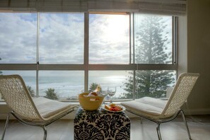 A room with two wicker chairs facing a large window overlooking a beach. A small table with a patterned tablecloth holds a bottle of champagne, a glass, and a plate with snacks. - Vue Devant (Cape Town)