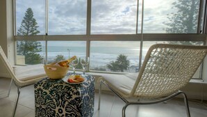 A room with a large window overlooking the ocean, a wicker chair, a small table with a bowl and glasses, and a patterned ottoman. - Vue Devant (Cape Town)
