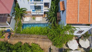 An aerial view of a hotel with a swimming pool, palm trees, and a thatched roof structure.