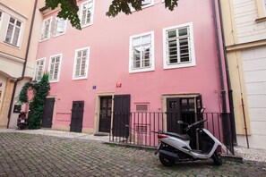 Un bâtiment rose aux fenêtres et aux portes blanches, un scooter garé devant, et une rue pavée.