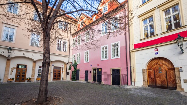 A cobblestone street with buildings, including a pink building with a red awning and a white building with a wooden door. - Tyn Yard Residence (Prague)