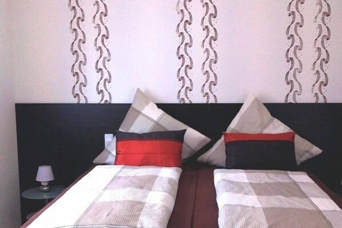 Una cama doble con un edredón a cuadros y dos almohadas, una roja y otra negra, en una habitación con papel tapiz estampado y cabecera oscura.
