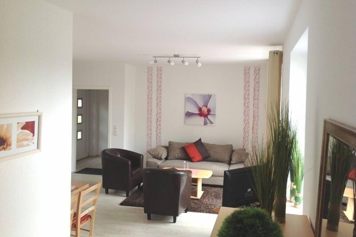 Una sala de estar moderna con sofá, sillones, una mesa de centro y televisor. Hay un cuadro en la pared y una planta en la esquina.