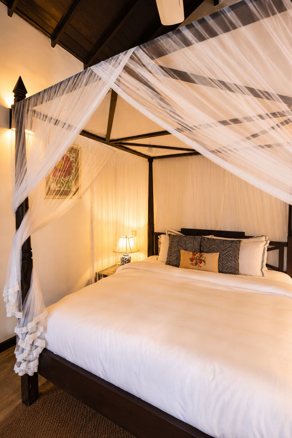 A four-poster canopy bed with white linens, a nightstand with a lamp, and a wall-mounted picture.