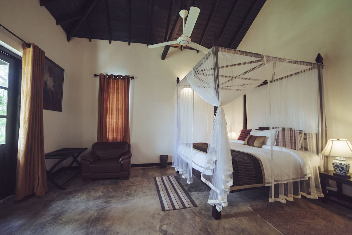 Ein Schlafzimmer mit einem Himmelbett, einem Deckenventilator, einem Sessel und einem Nachttisch mit Lampe.