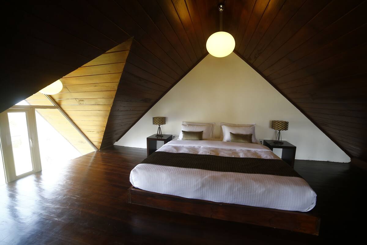 A bedroom with a sloped ceiling, a bed with white bedding, two bedside tables with lamps, and a view of the outside through a window.