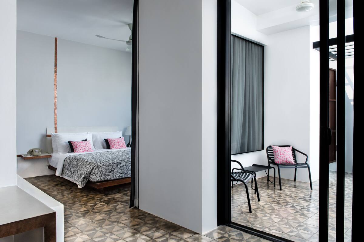 A modern hotel room with a bed, a small table, and a chair. There is a window with curtains and a view of the outside.