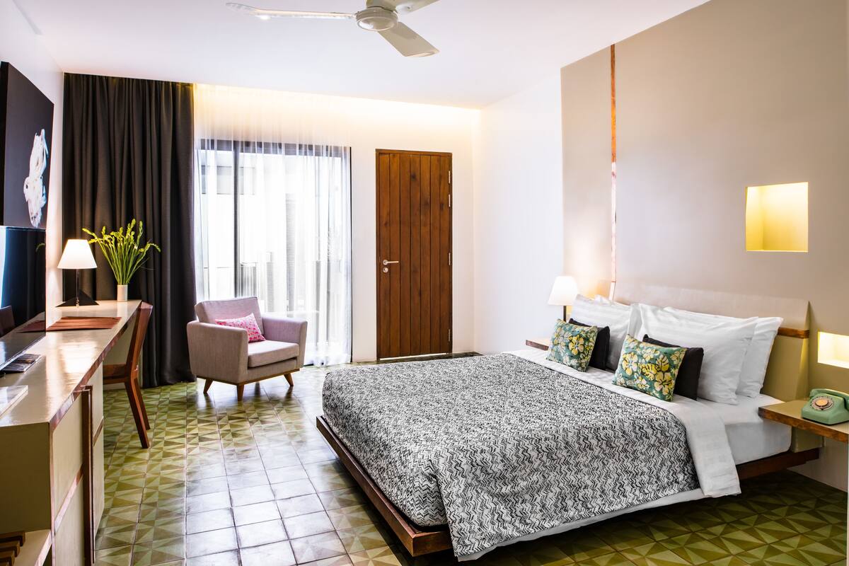 A hotel room with a bed, bedside table, lamp, chair, and a desk with a lamp.