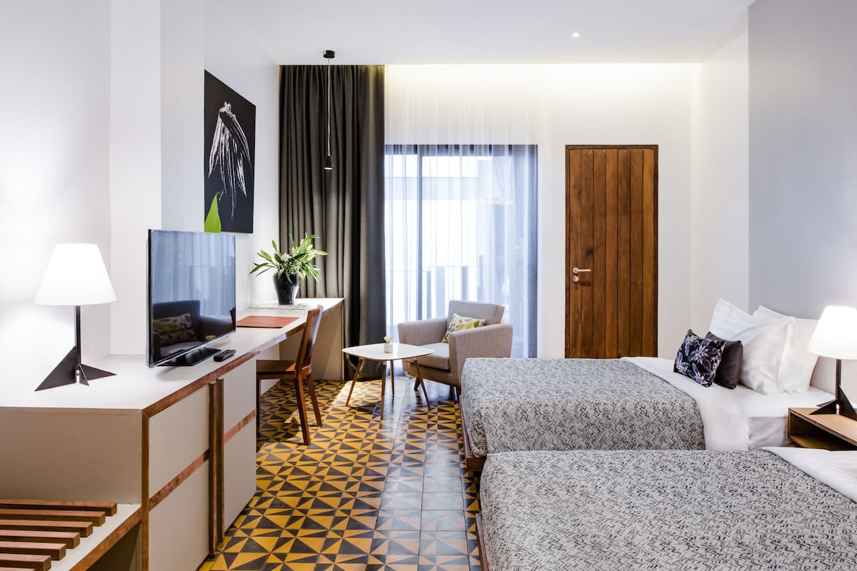 A hotel room with a bed, a desk with a TV, a chair, a small table, and a plant.
