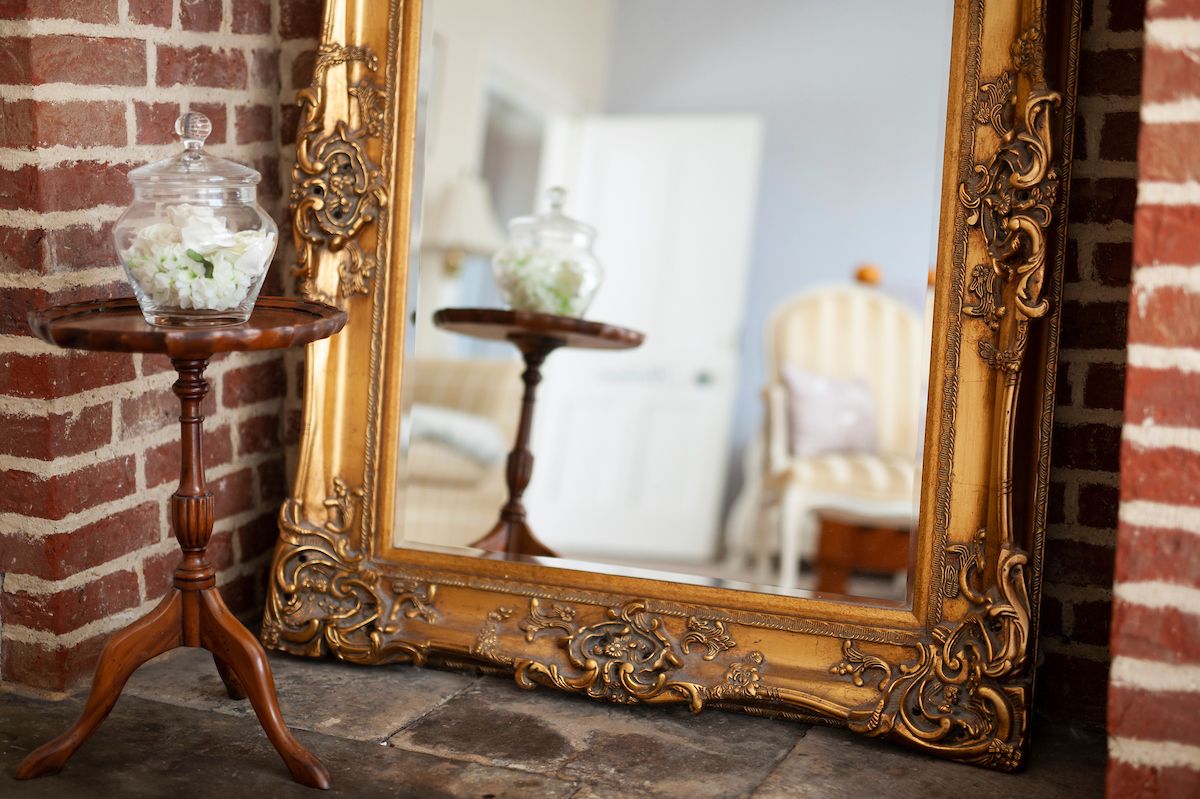 A mirror with an ornate gold frame, a small wooden table with a glass jar of white flowers, and a brick wall in the background.