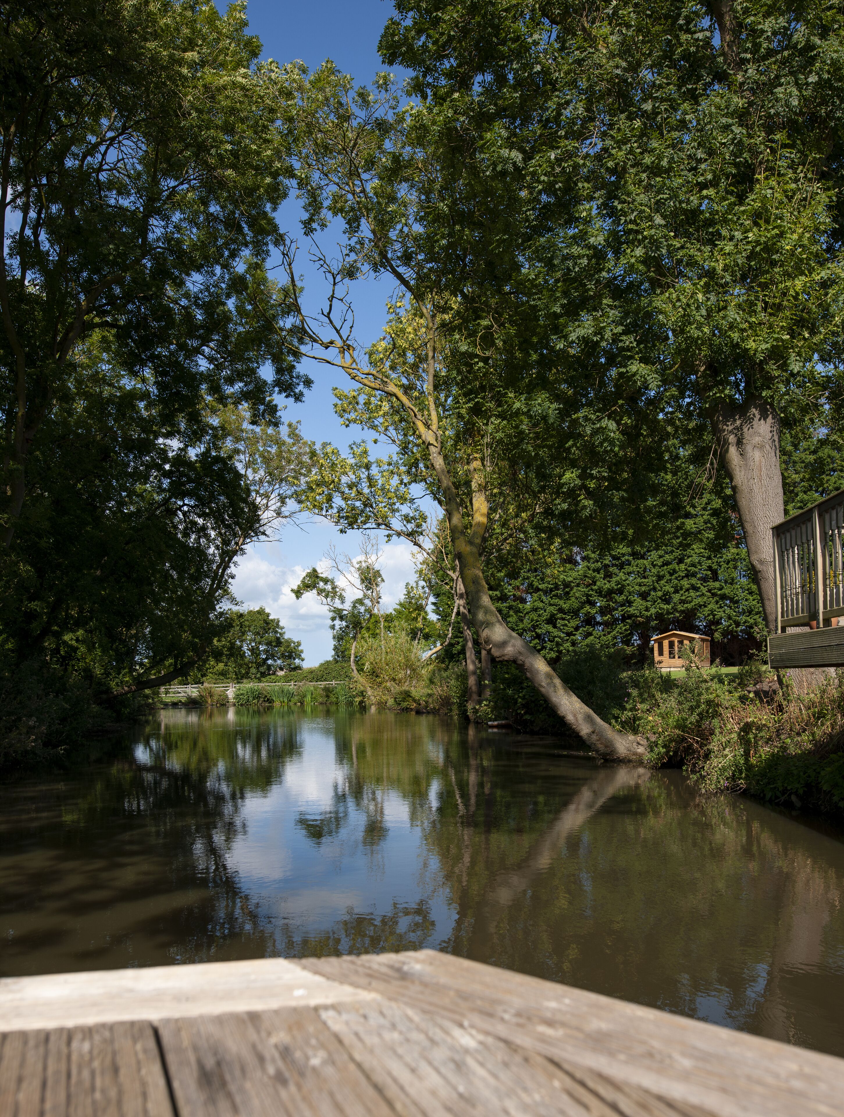 A wooden dock extending into a calm river, surrounded by lush greenery and a small wooden structure on the opposite bank.
