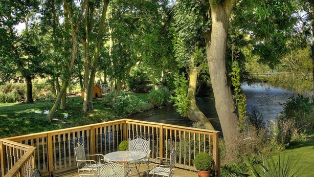 Une terrasse en bois, agréée de mobilier d’extérieur, entourée d’une végétation luxuriante et bordée d’un étang.