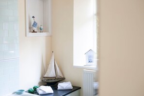 Un baño con pared de azulejos blancos, un pequeño modelo de velero, una ventana con vista a una casa y un perchero con toallas dobladas.