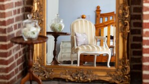 Une pièce dotée d’un grand miroir décoratif qui reflète un fauteuil à rayures, une petite table surmontée d’un récipient en verre et une chaise en bois.