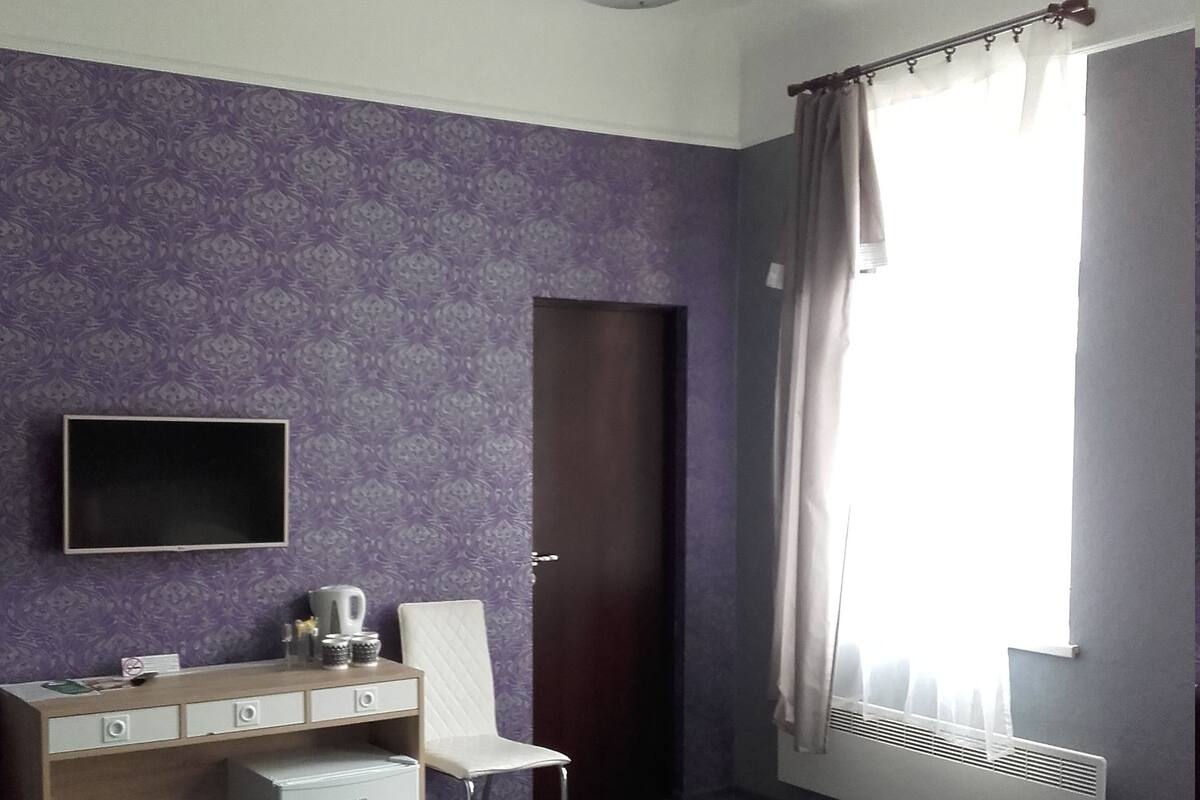 A hotel room with a bed, a desk with a chair, a television, and a window with curtains.