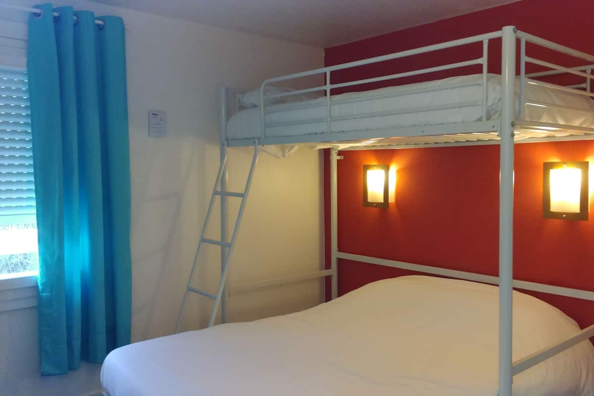 Una cama litera con escalera, una ventana con cortinas azules y dos luces fijadas a la pared.
