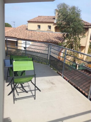 Een balkon met een groene tafel en stoelen, een metalen hekwerk en uitzicht op een gebouw met een pannendak.