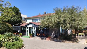 Un hotel con vestíbulo cubierto, mesas al aire libre y un jardín con diversas plantas.