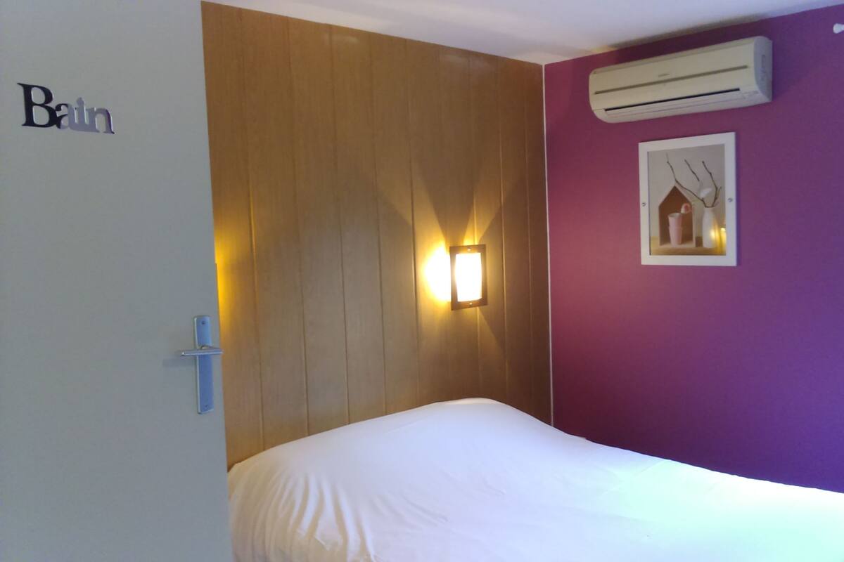 Una habitación con una cama, aire acondicionado de pared, una lámpara de pared y un cuadro enmarcado.