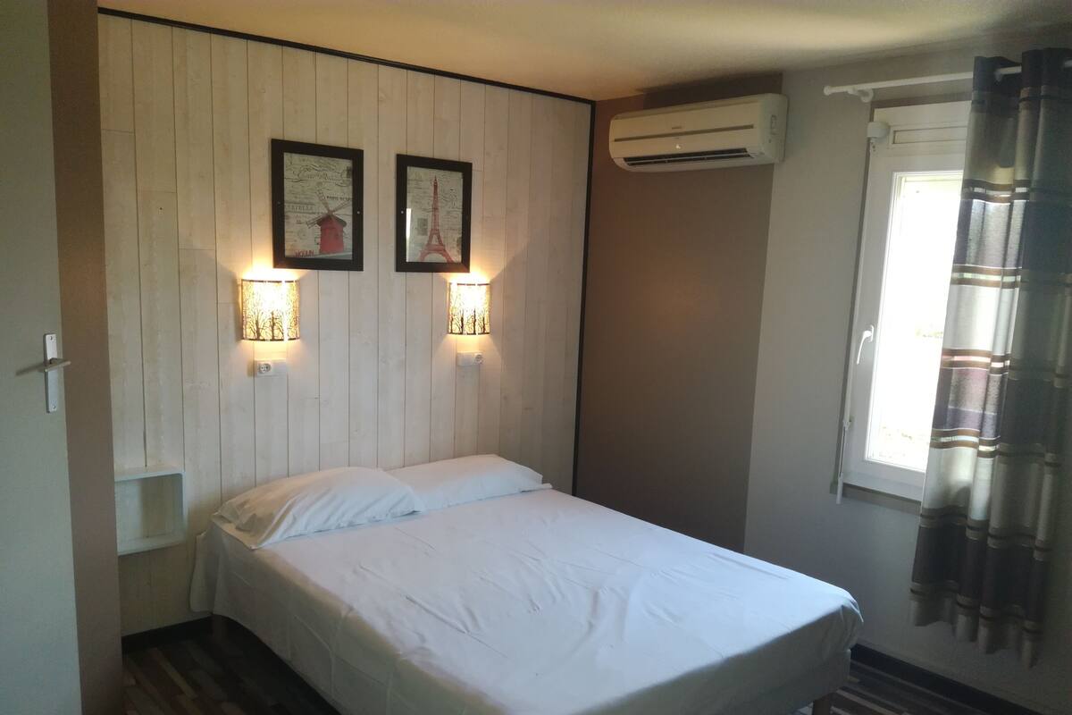 Habitación individual con pared de paneles de madera, dos cuadros enmarcados, un equipo de aire acondicionado y una ventana con cortinas.