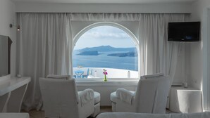 Habitación de hotel con un amplio ventanal de arco, cortinas blancas, dos sillas blancas, una mesita, un televisor de pantalla plana y vistas al mar y a las islas.