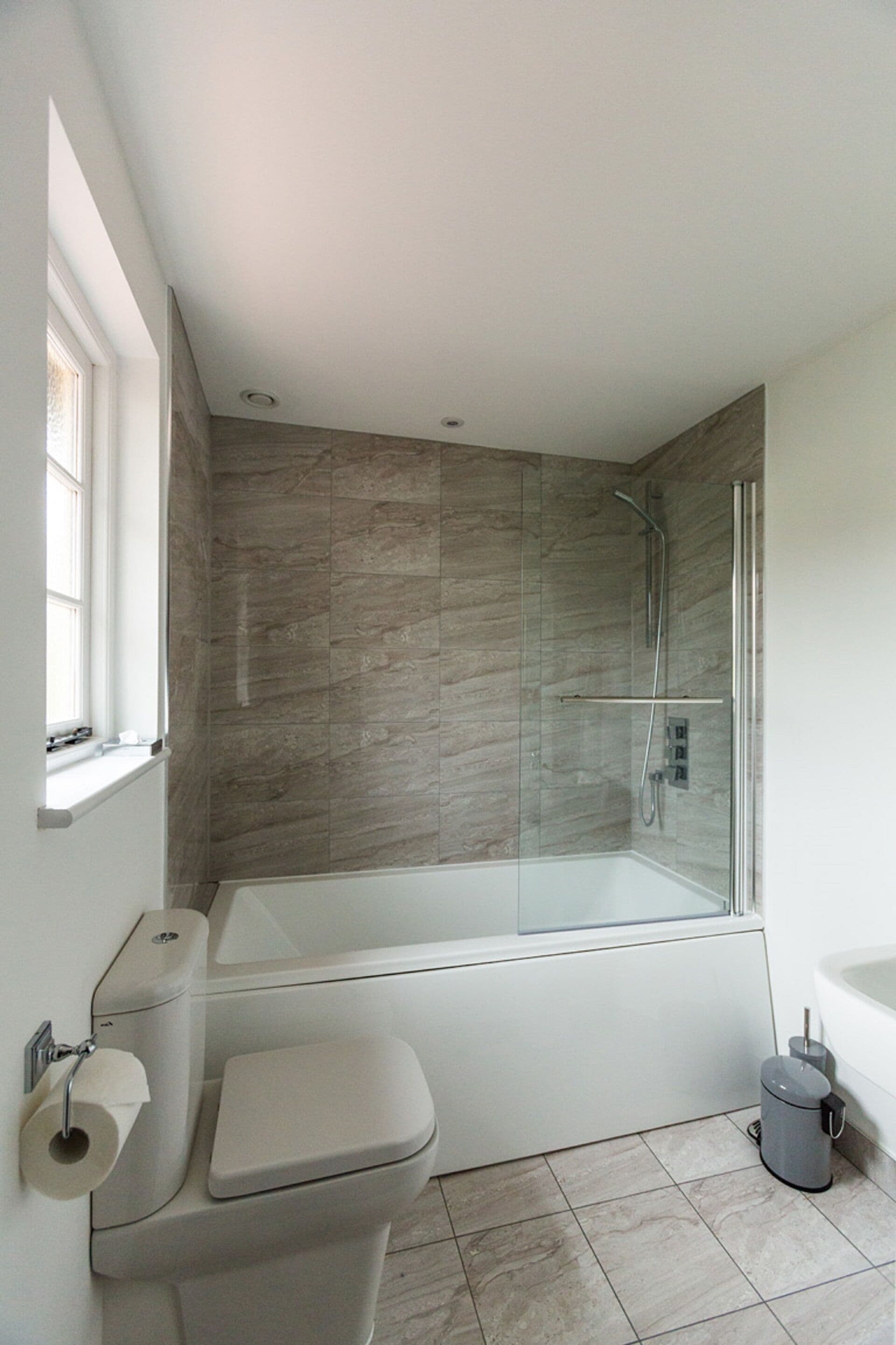 A modern bathroom with a glass-enclosed shower, a white bathtub, and a toilet with a paper towel holder.