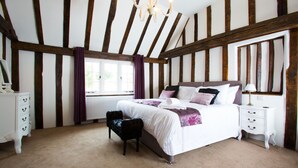 A bedroom with exposed wooden beams, a large bed with white bedding and purple accents, a nightstand, a dresser, and a window with curtains. - The Harvard Inn (Ingatestone)