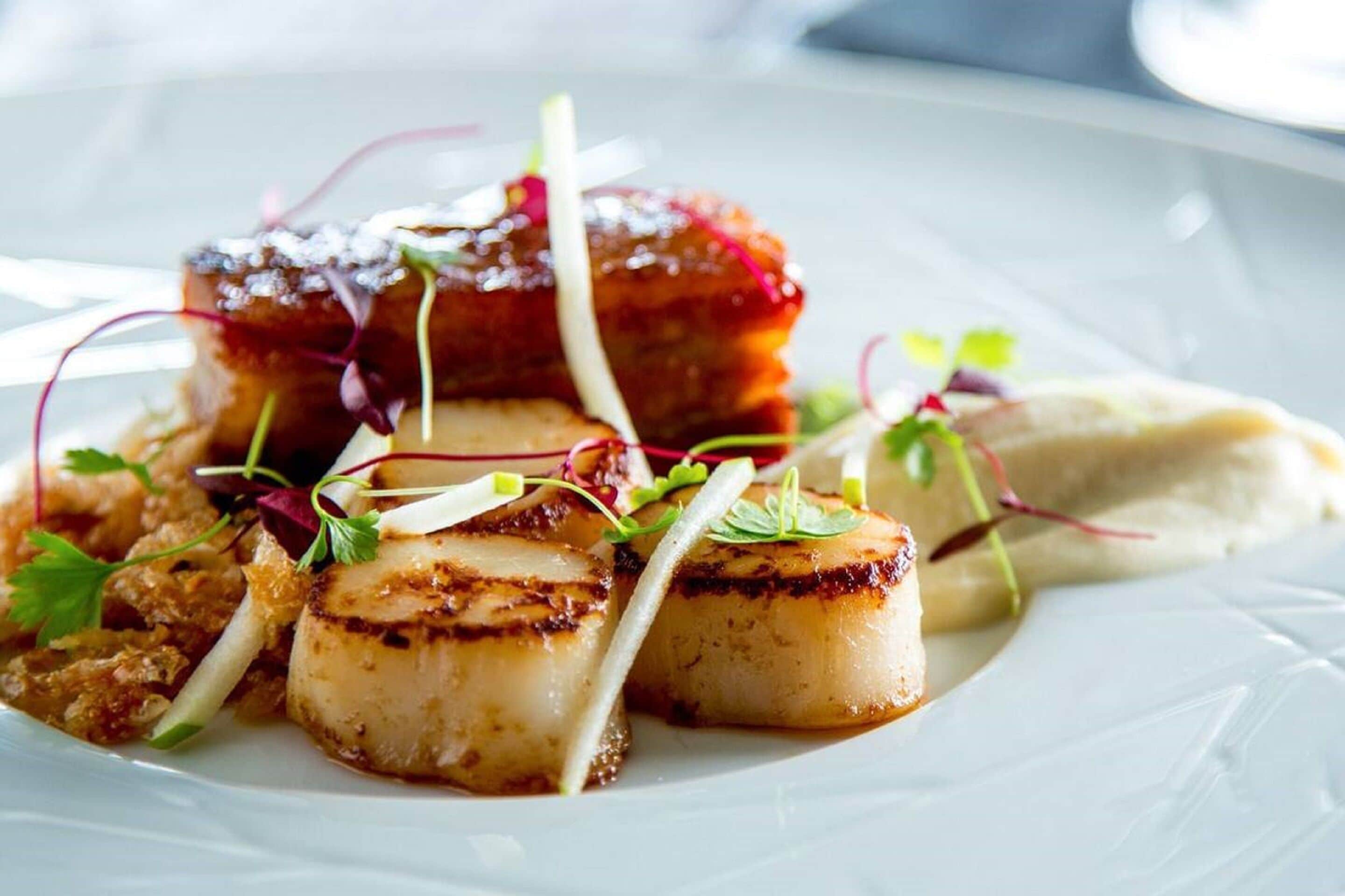 Un plato de vieiras a la plancha, adornado con microgreens y verduras en rodajas finas.
