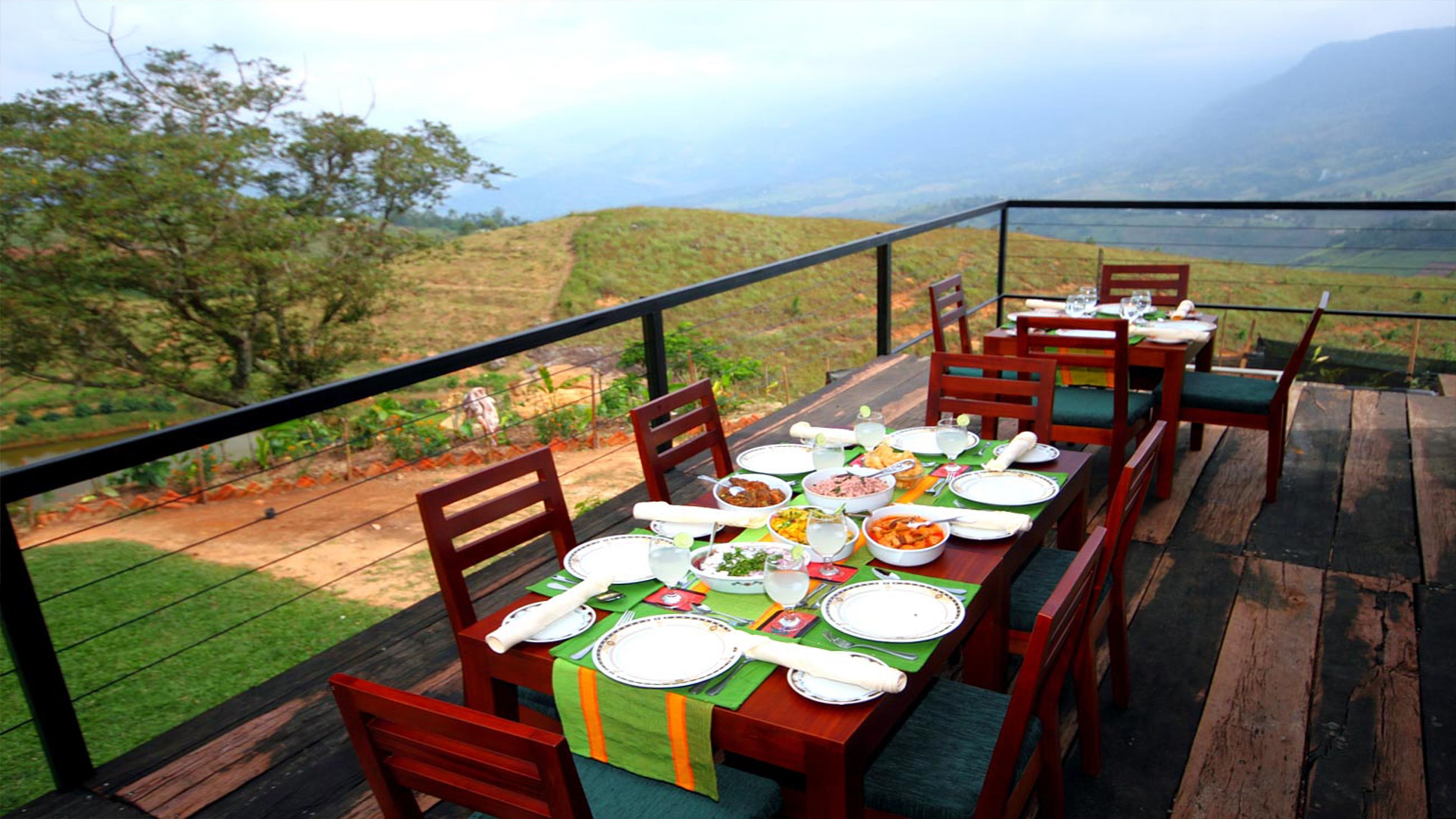Une longue table à manger en bois, dressée avec des assiettes, des bols et des couverts, entourée de chaises, sur une terrasse extérieure offrant une vue magnifique.