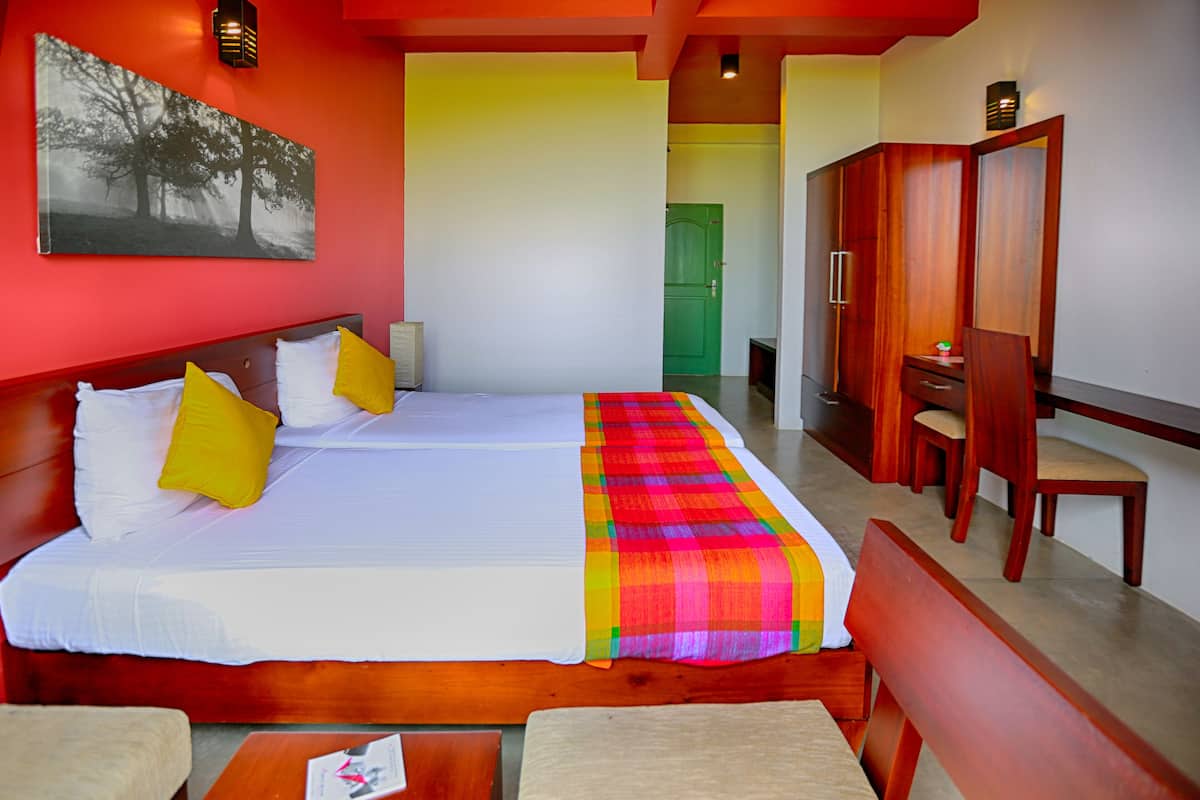 A hotel room with a large bed, two pillows, a colorful blanket, a wooden headboard, a desk, and a chair.
