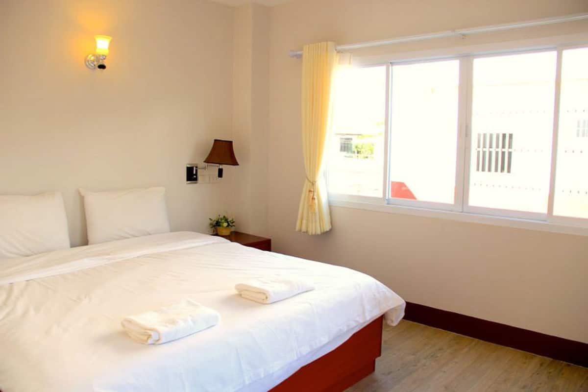 A neatly made bed with white linens and two towels, a bedside lamp, a small table with a plant, and a window with curtains.