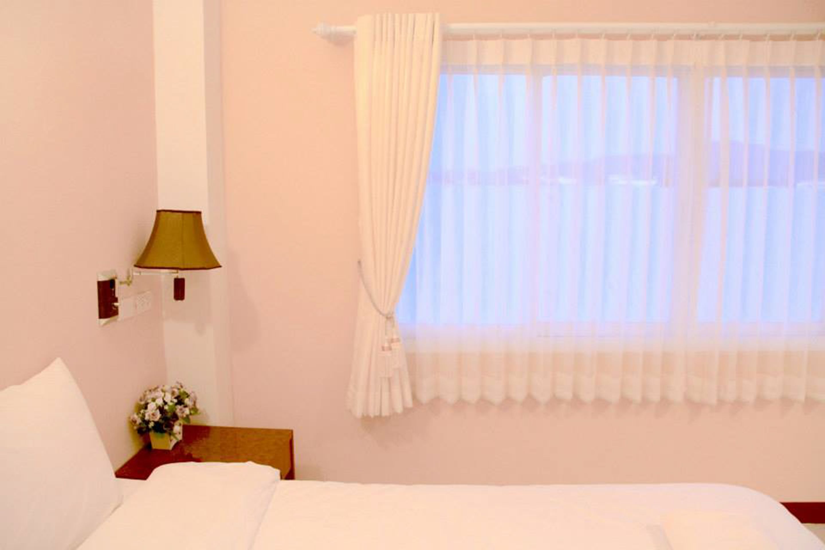 A hotel room with a bed, a nightstand with a lamp, a window with sheer curtains, and a wall-mounted light switch.