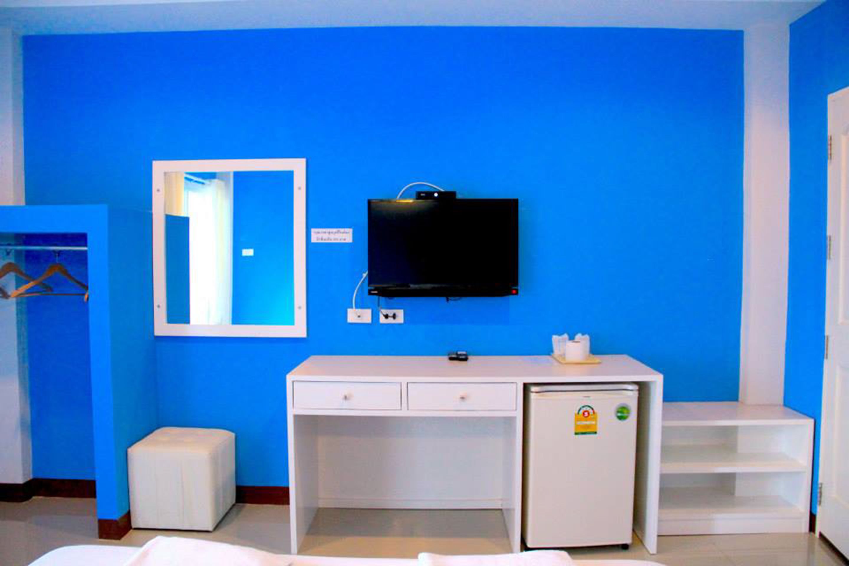 A room with a blue wall, a white desk with drawers, a small refrigerator, a television mounted on the wall, and a mirror above the desk.