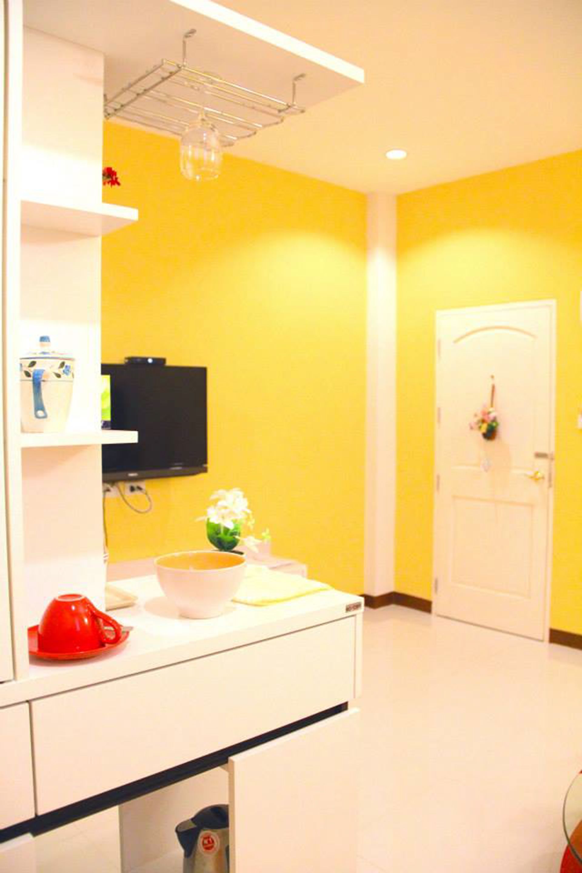 A bright yellow room with a white counter, a television mounted on the wall, and a door with a flower wreath.