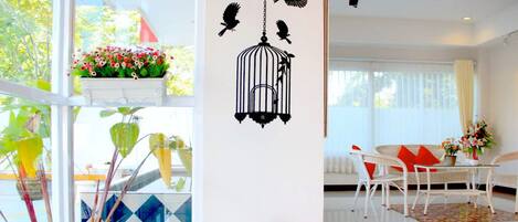 Una habitación luminosa con pared blanca, una jaula de pájaro decorativa colgante, una figurita pequeña y un área de comedor con sillas y mesa blancos.