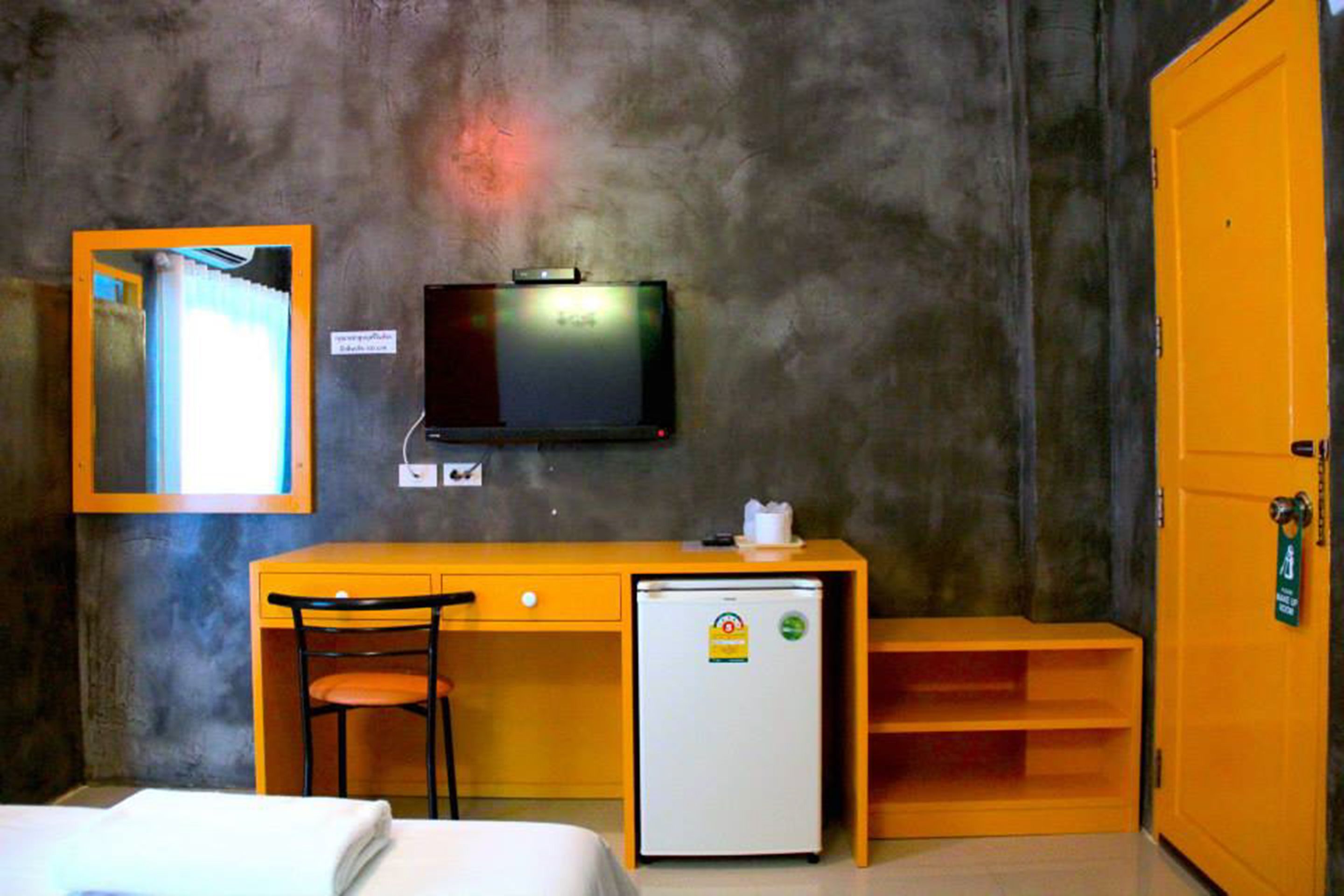 A compact hotel room with a bed, a desk with a chair, a small refrigerator, a television mounted on the wall, and a door with a sign.