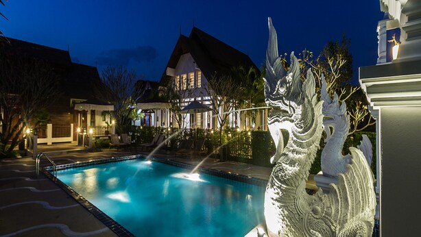 Una piscina con una escultura decorativa, una casa con techo de paja y un área exterior bien iluminada.