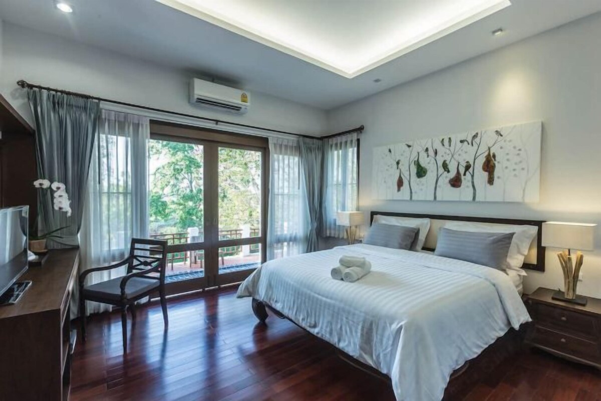 Un dormitorio con una cama grande, un escritorio y una silla. Hay una ventana con cortinas, un cuadro en la pared y un aire acondicionado.