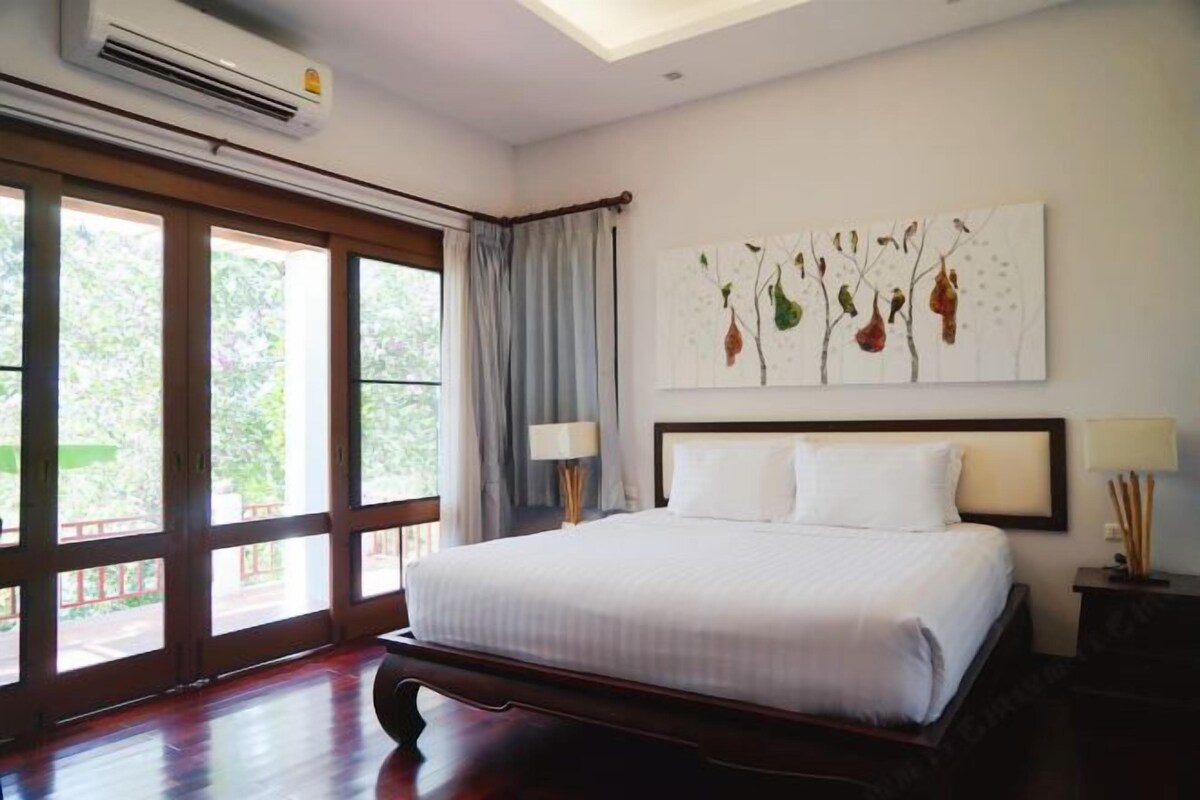 A hotel room with a large bed, a ceiling fan, a window with curtains, and a painting on the wall.