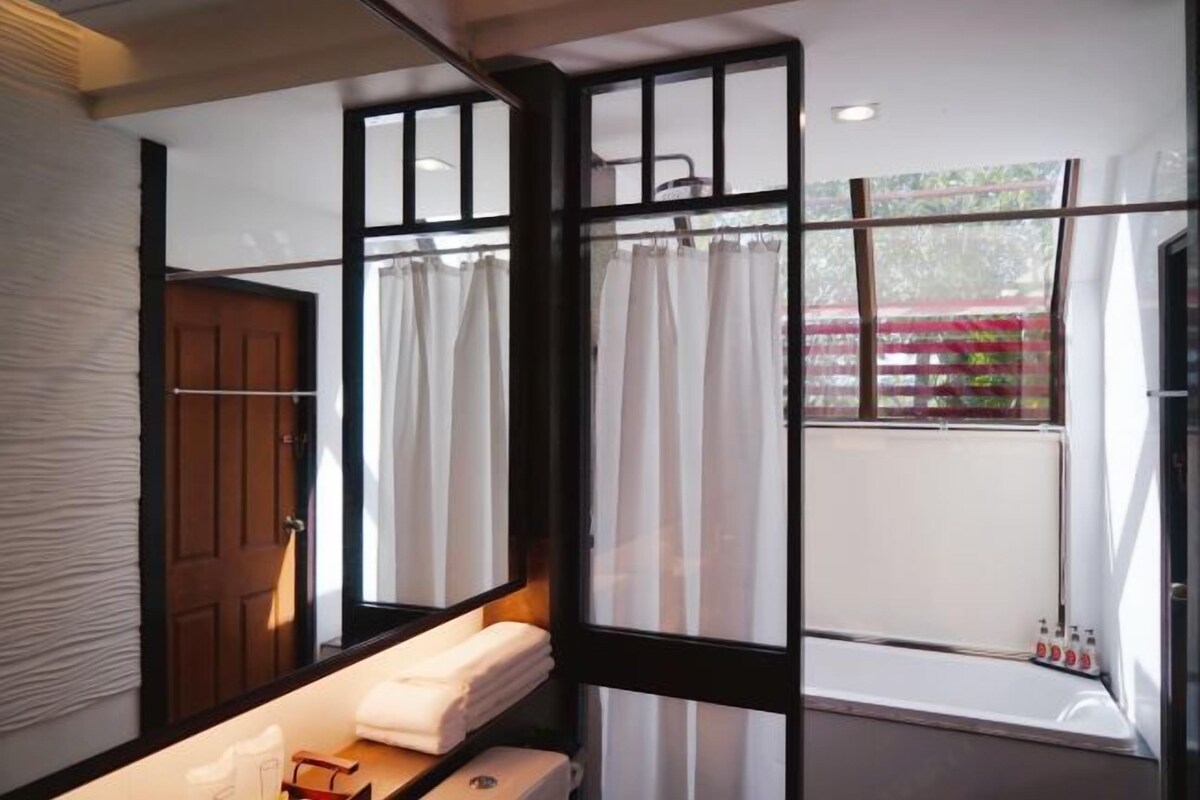Um banheiro com porta de madeira, janela com cortinas e prateleira com toalhas.