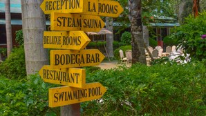 Placa de indicadores com direções para diversas comodidades do hotel, incluindo recepção, quartos, sauna a vapor, quartos deluxe, bar bodega, restaurante e piscina.