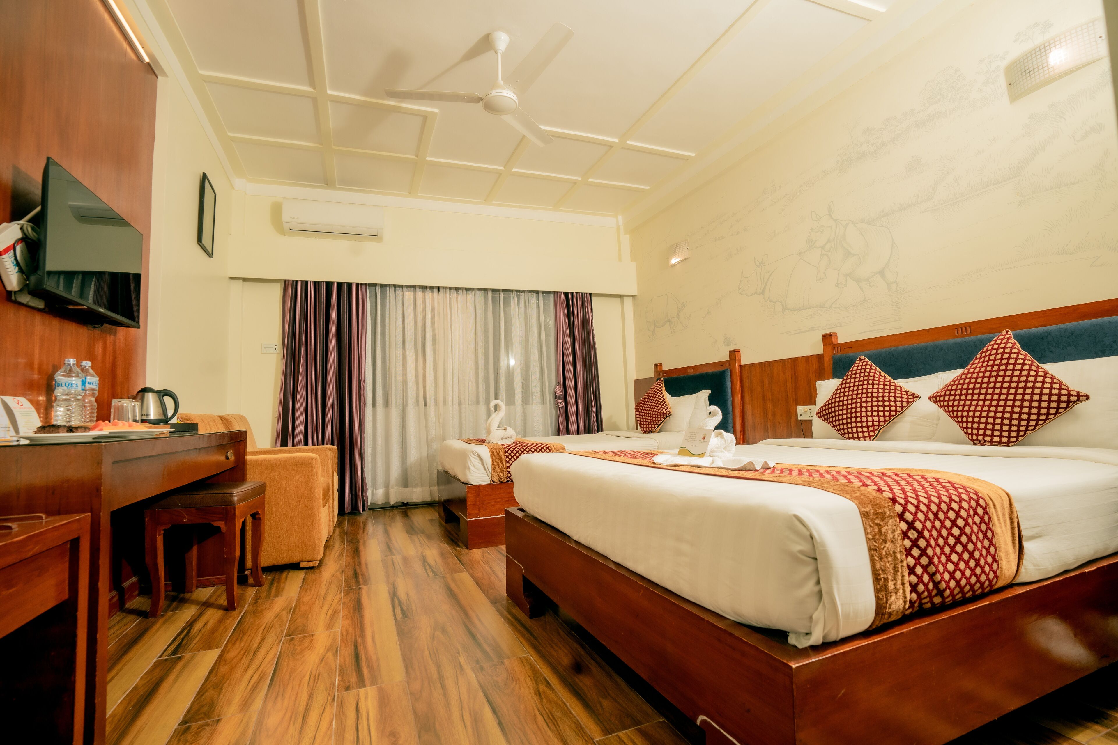 Een hotelkamer met een houten bureau, een bed met gestreepte kussens, een plafondventilator en een groot raam met gordijnen.